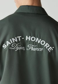 Dos d'une veste vert foncé avec texte blanc brodé « SAINT-HONORÉ Dijon, France » visible près du col.