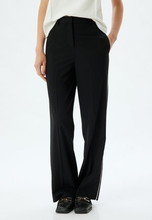 Pantalons noirs à jambes larges avec une texture lisse, présentant une coupe ajustée et des rayures blanches sur les côtés. Portés avec des chaussures noires texturées.