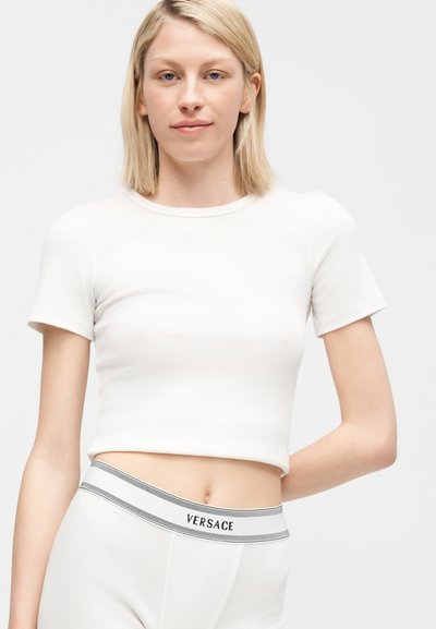 Jeune femme aux cheveux blonds portant un t-shirt court blanc et un pantalon blanc avec une ceinture avec le logo Versace, posant devant un fond uni.