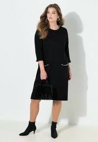 Schwarzes knieumspielendes Kleid mit dreiviertel Ärmeln, verziert mit Perlenakzenten an Taschen und Manschetten. Kombiniert mit einer schwarzen Handtasche mit Fransen.