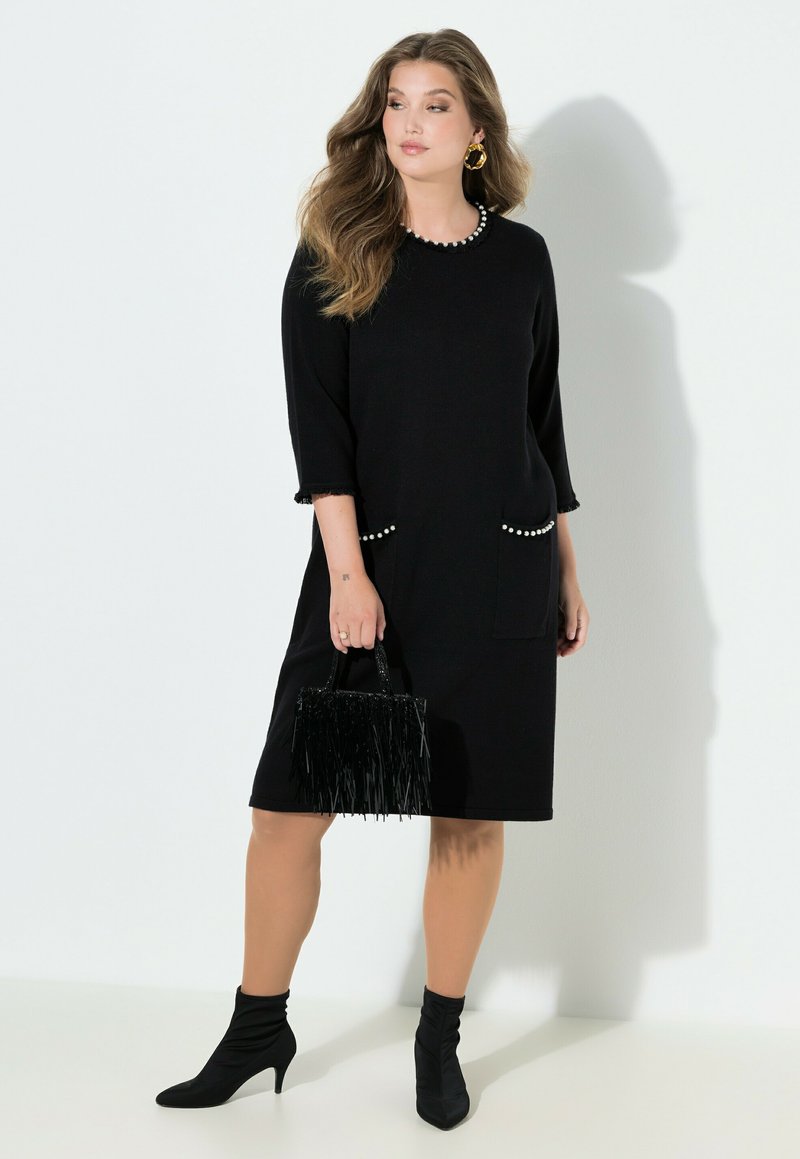 Schwarzes knieumspielendes Kleid mit dreiviertel Ärmeln, verziert mit Perlenakzenten an Taschen und Manschetten. Kombiniert mit einer schwarzen Handtasche mit Fransen.