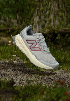 Chaussure de running New Balance grise avec logo rouge et semelle blanche flottant au-dessus de gravier et de verdure en plein air.