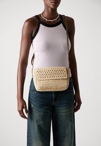 Borsa all'uncinetto realizzata con fibre naturali, di colore beige con un pattern intrecciato e testurizzato. Presenta una chiusura a patta e una lunga tracolla regolabile.