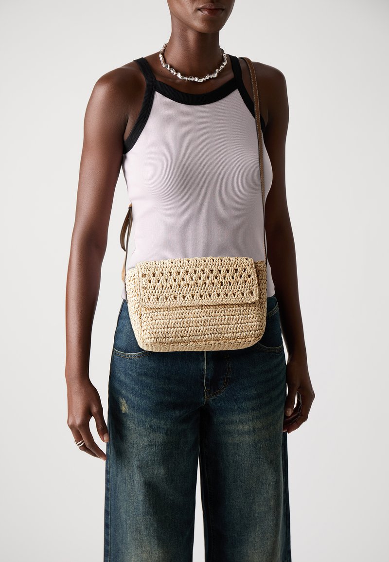 Borsa all'uncinetto realizzata con fibre naturali, di colore beige con un pattern intrecciato e testurizzato. Presenta una chiusura a patta e una lunga tracolla regolabile.