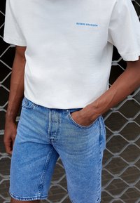 Camiseta de algodón blanca con "QUEENS BOROUGH" impreso en azul, combinada con pantalones cortos de mezclilla azul claro. Textura notable y ajuste clásico.