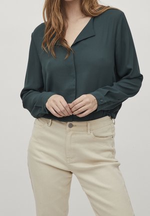 Kvinde iført en mørkegrøn bluse med lange ærmer, stoppet ned i højtaljede beige bukser, hænderne foldet ved taljen.