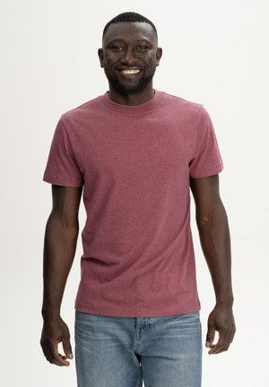 Lachende man met een effen bordeauxrood T-shirt met korte mouwen en blauwe spijkerbroek, staand tegen een lichte neutrale achtergrond.