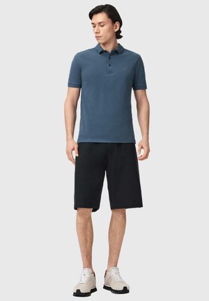 Mężczyzna stojący, ubrany w niebieską koszulkę polo z krótkim rękawem, czarne szorty za kolano i beżowe trampki, patrzący w lewo.