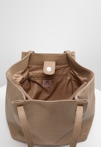 Bolso tote de cuero beige con interior de tela, cierre superior con cremallera, bolsillo interior con cremallera y dos asas para el hombro. Incluye un botón de presión.