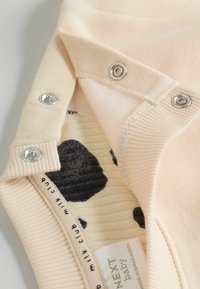 Bodysuit para bebé color durazno con cierres a presión de metal. El interior presenta un patrón de manchas en negro y crema sobre tela acanalada. Etiqueta del fabricante visible.