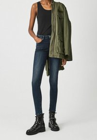 Hubené tmavě modré džíny, černý tílko a olivově zelená vojenská bunda, doplněné černými šněrovacími bojovými botami.