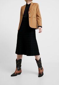 Blazer marron à deux boutons, blouse noire, jupe plissée noire, et bottes de cowboy en cuir noir et marron avec des accents étoilés.