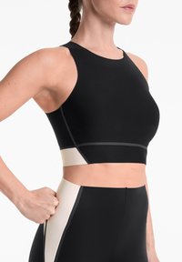 Soutien-gorge de sport noir avec un col montant et des panneaux beiges en contraste. Texture lisse, couture minimale, et design ajusté mettant en valeur la taille.