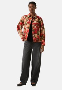 Jacke mit Blumenmuster in Rot, Creme und Grün aus Baumwolle, mit Kragen und Brusttaschen, kombiniert mit schwarzen, weit geschnittenen Jeans und schwarzen Ballerinas.