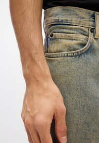 Világoskék farmernadrág enyhén kopott textúrával. Elülső zseb és kis címke található rajta. Közelkép egy kézről, amely a csípőn pihen.