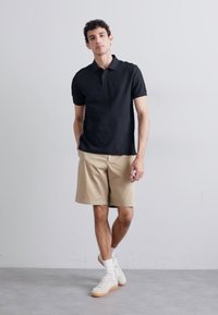 Melna polo krekls ar īsām piedurknēm, bēšas šortus un baltas potītes zeķes. Modelis valkā gaišas sporta kurpes. Vienkārša, ikdienišķa apģērba kombinācija.