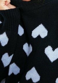Schwarzer Strickpullover mit hellblauen Herzmustern. Die Textur wirkt weich, mit einem hohen Kragen und gerippten Bündchen.