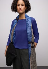 Pull bleu, écharpe à motifs bleus et bruns, pantalon gris et sac en daim noir. Les textures et couleurs remarquables renforcent l'attrait visuel de la tenue.