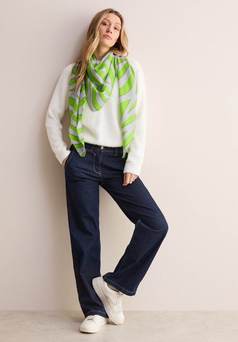 Hvid struktureret sweater, grøn og grå mønstret tørklæde, mørkeblå bukser med vidde og hvide sneakers, mod en neutral baggrund.