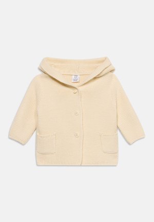 Gebreid babyvestje in crèmekleur met capuchon, drie knopen aan de voorkant en twee kleine voorzakjes, plat neergelegd op een witte achtergrond.