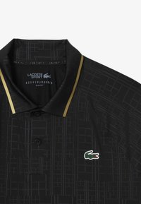 Svart pikétröja med ett geometriskt mönster, guldaccenter på kragen och en liten grön krokodil-logotyp. Slim fit-design och knappslå.