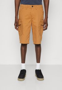 Knielange Cargo-Shorts aus senfgelben Baumwolle mit zwei großen Seitentaschen, einem Knopfverschluss und Gürtelschlaufen. Getragen mit schwarzen Sneakers.