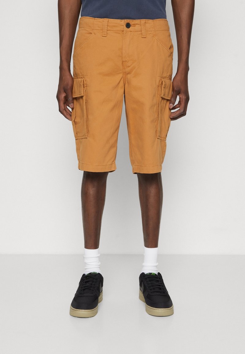 Knielange Cargo-Shorts aus senfgelben Baumwolle mit zwei großen Seitentaschen, einem Knopfverschluss und Gürtelschlaufen. Getragen mit schwarzen Sneakers.