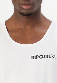 Tricou alb fără mâneci, cu un guler rotund și logo-ul negru "RIPCURL" cu un design de val pe față, textura din bumbac neted.