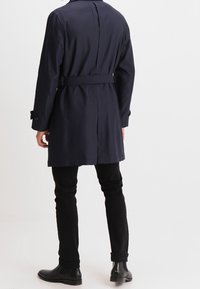 Granatowy płaszcz trencz z paskiem w talii, zapinany na guziki przy mankietach, o gładkiej fakturze; zestawiony z dopasowanymi czarnymi spodniami i botkami za kostkę.