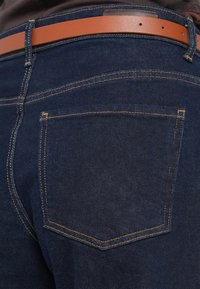 Tamno plave traperice imaju svijetlosmeđi kožni remen. Traperice imaju žute šavove i klasičan dizajn s pet džepova uz suptilnu teksturu.