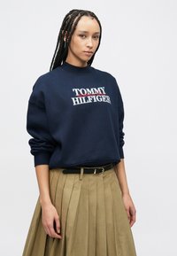 Μακρυμάνικο μπλε σκούρο φούτερ με το λογότυπο «TOMMY HILFIGER» σε αντιθετικό λευκό και κόκκινο. Διαθέτει στρογγυλή λαιμόκοψη και άνετη εφαρμογή, συνδυασμένο με φούστα με πιέτες.