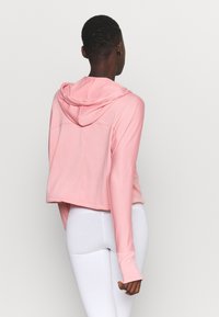 Sweat à capuche court rose avec capuche, manches longues et passants pour les pouces. Texture lisse et design simple. Porté avec des leggings blancs.