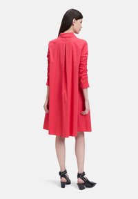 Robe chemise rouge à manches longues et détail plissé dans le dos. Fabriquée en tissu lisse, elle présente des manches élastiquées et est assortie à des sandales à talons noires.