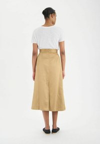 Jupe A-line beige jusqu'aux genoux en tissu lisse, avec une taille haute et une fermeture éclair discrète au dos. Associée à un simple T-shirt blanc.