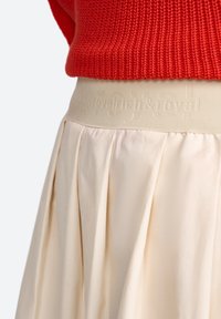 Jupe plissée en crème avec une taille élastique texturée arborant une marque en relief. Le tissu semble lisse et léger.