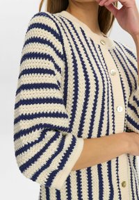 Strickjacke mit cremefarbenen und marineblauen horizontalen Streifen, ausgestattet mit einem runden Ausschnitt und Perlenknöpfen an der Vorderseite. Weicher, strukturierter Stoff.