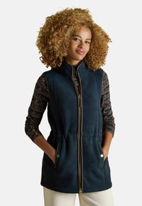 Sötétkék bolyhos magasszájas mellény, cipzáros záródással, két elülső zsebbel és rugalmas derékkal; mintás, hosszú ujjú felsőre van felvéve.