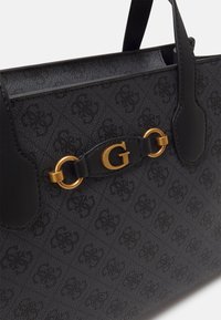 Musta kuviollinen käsilaukku, jossa on teksturoitu pinta, kultaiset yksityiskohdat ja suuri "G"-koriste edessä.
