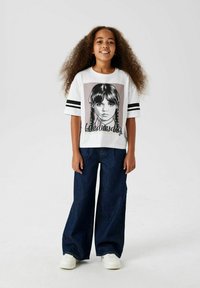 Vit t-shirt med grafiktryck av en tjej med flätor, svarta ränder på ärmarna, i kombination med vida blå denimjeans och vita sneakers.