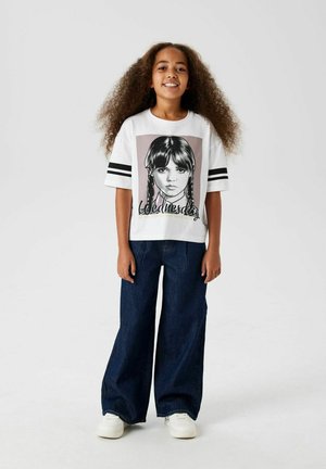 T-shirt bianca con stampa grafica di una ragazza con trecce, strisce nere sulle maniche, abbinata a jeans blu a gamba larga e sneakers bianche.