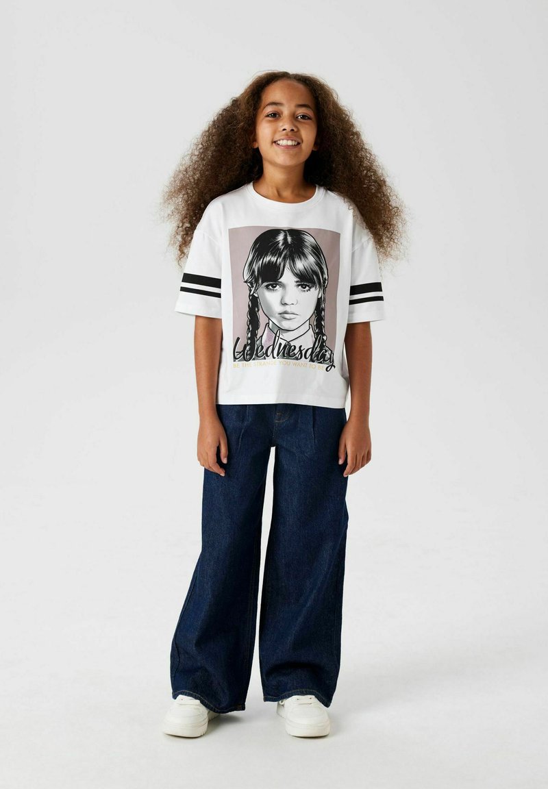 Vit t-shirt med grafiktryck av en tjej med flätor, svarta ränder på ärmarna, i kombination med vida blå denimjeans och vita sneakers.