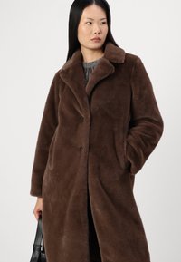 Cappotto in pelliccia sintetica marrone con colletto a punte, bottoni frontali e tasche laterali. Tessuto texture e design di lunghezza media.