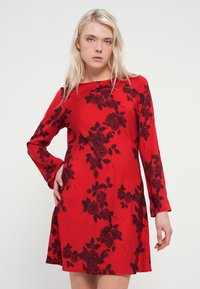 Rdeča obleka z dolgimi rokavi z črnim cvetličnim vzorcem. Tkanina je gladka in se nežno razširi od pasu naprej.