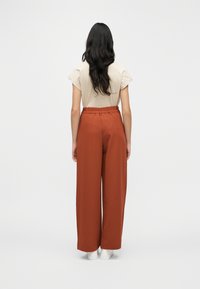 Μπεζ τοπ με κοντά μανίκια και βολάν στους ώμους, συνδυασμένο με ψηλόμεσα παντελόνια σε φαρδιά γραμμή σε χρώμα σκουριάς και λευκά αθλητικά παπούτσια.