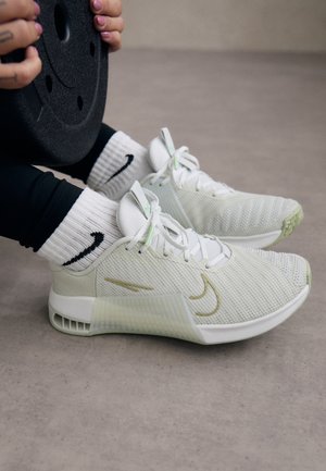 White athletic shoes with mesh texture and green accents worn with white Nike socks, person holding a black weight plate.