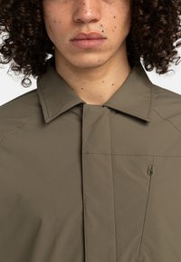 Olive green, lightweight jacket with a pointed collar, subtle stitching details, and a small front pocket with a zipper pull.