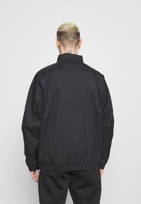 Osoba s krátkými bělenými vlasy a tetováním na krku, oblečená v černé bundě a černých kalhotách, viděná zezadu na bílém pozadí.