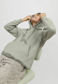 Sudadera verde claro con bolsillo canguro, capucha con cordón y logotipo sutilmente embosado. Lleva puesta sobre pantalones grises claros con textura, sentado en una silla.