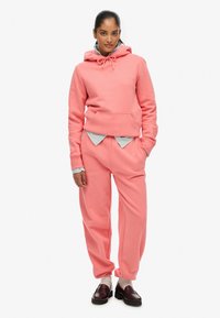 Conjunto de sudadera y pantalones de chándal en suave tela rosa. Cuenta con capucha, bolsillo canguro y puños acanalados. Se lleva con una camiseta ligera de color azul claro debajo.