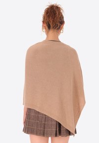 Poncho beige realizzato in materiale morbido, con un design drappeggiato, apertura frontale e finiture senza cuciture. Indossato sopra una gonna marrone a fantasia.